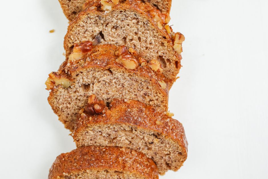 A close-up shot of a golden-brown banana nut bread loaf with a glossy white glaze dripping down the sides, topped with chopped toasted pecans.