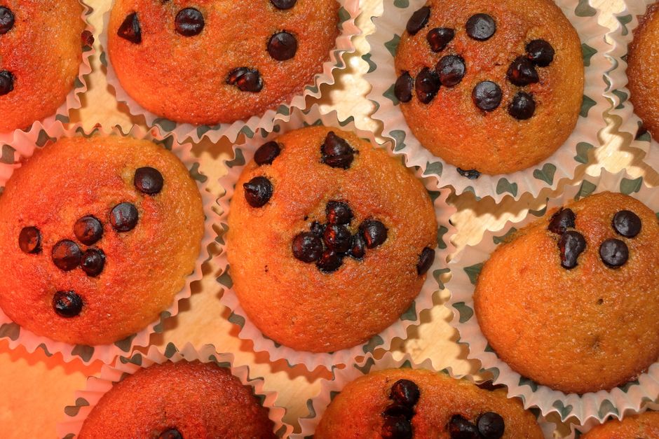 A close-up shot of freshly baked banana bread chocolate chip muffins in a muffin tin, with melted chocolate chips on top and steam gently rising.