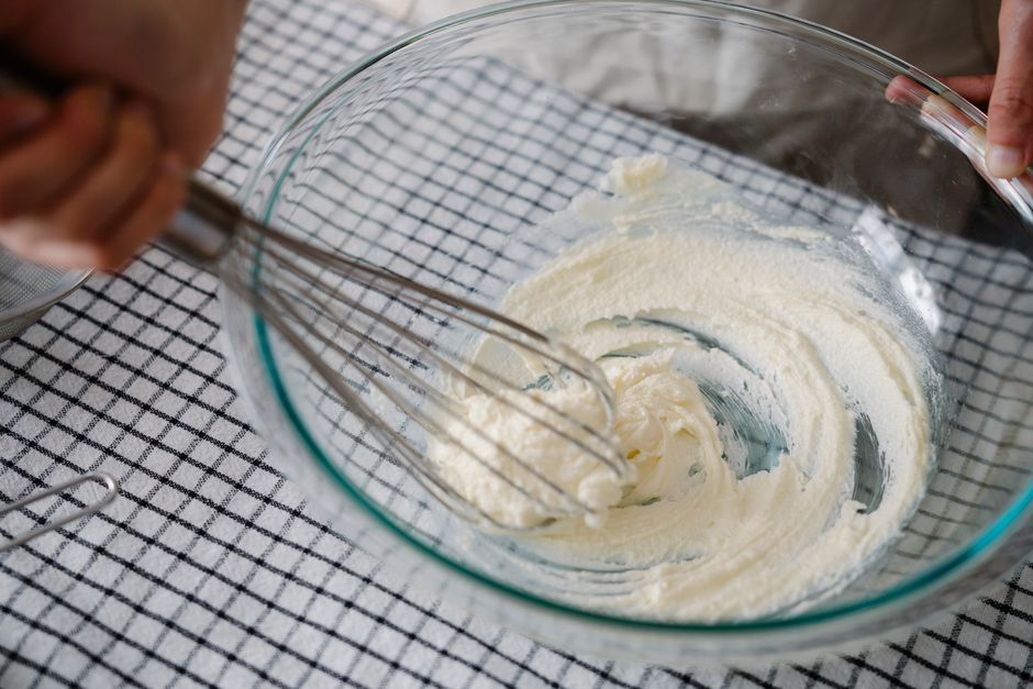 A glass mixing bowl showing the smooth, creamy glaze being whisked, with a bottle of pure maple syrup and a bowl of walnuts in the background.