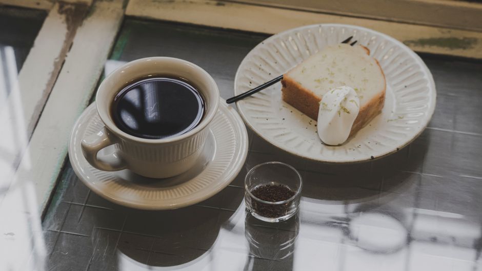 A slice of glazed banana nut bread served on a ceramic plate next to a cup of hot black coffee.
