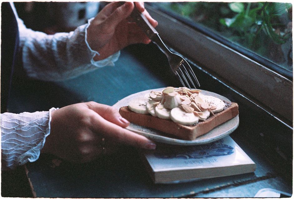 A slice of iced banana bread served on a white plate with a fork, with a cup of coffee in the background