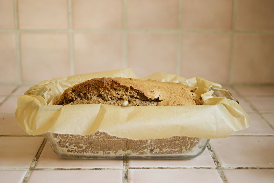 A warm, freshly baked banana loaf sitting next to a modern bread maker on a rustic kitchen counter